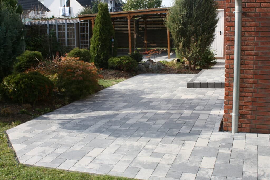 Patio moderne en pavés gris clair et foncé, avec marches. Jardin paysager, pergola bois, façade en briques et descente pluviale.