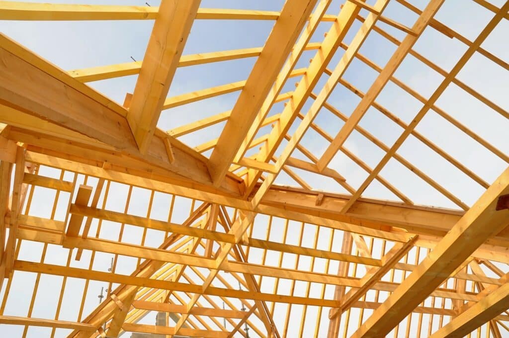 Structure de charpente en bois d'un toit en construction vue d'en bas contre un ciel lumineux. Poutres et chevrons forment un motif complexe.