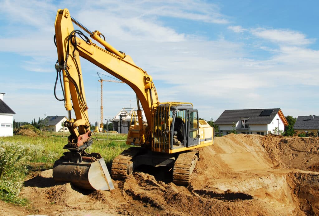 Pelleteuse jaune creusant un chantier de construction avec des maisons neuves et une grue sous un ciel nuageux.