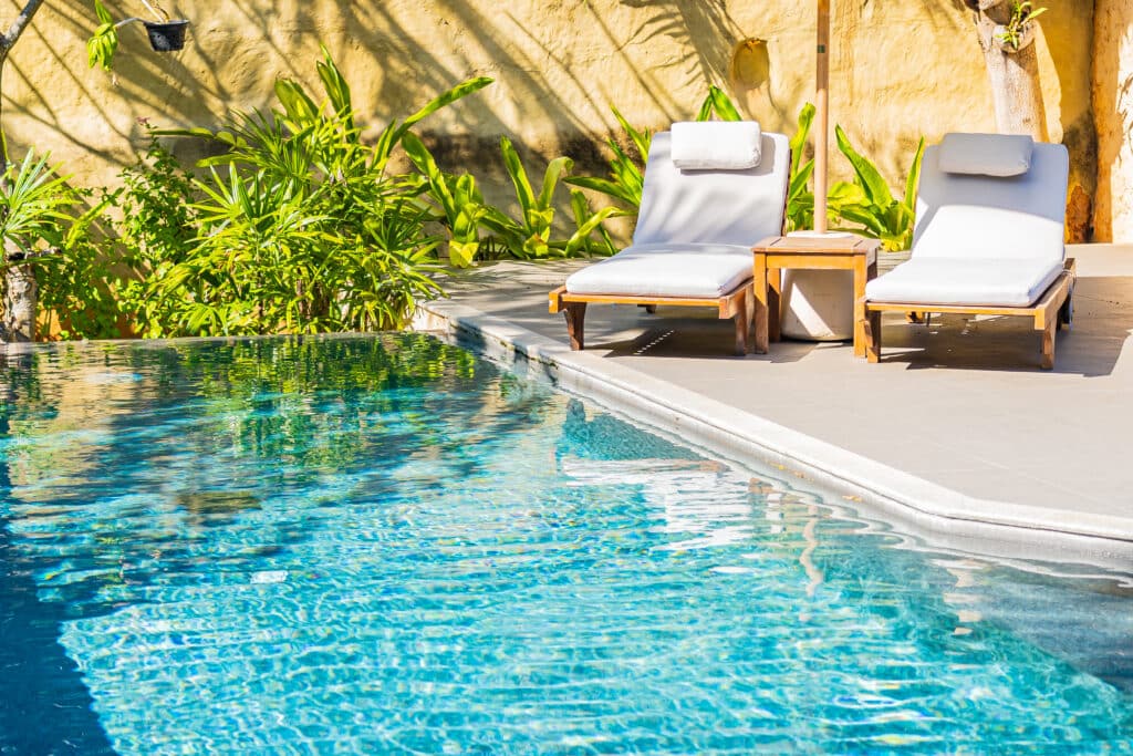 Piscine d'hôtel avec eau turquoise, deux chaises longues blanches sur une terrasse ensoleillée. Verdure luxuriante et mur texturé en arrière-plan.