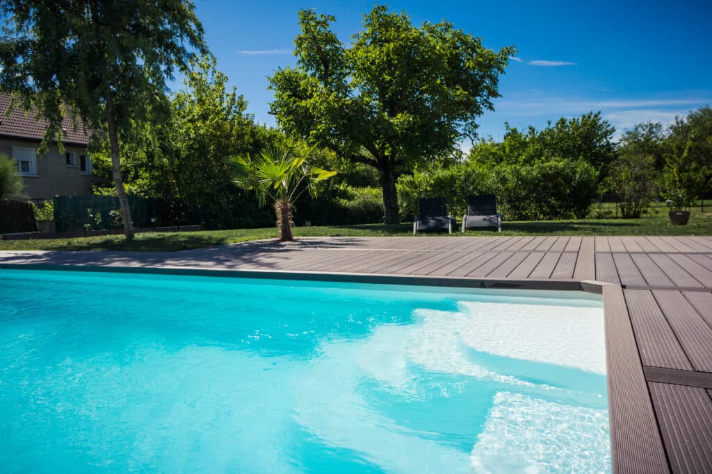 Piscine privée moderne avec eau turquoise et escalier, entourée d'une terrasse en bois et d'un jardin luxuriant ensoleillé.
